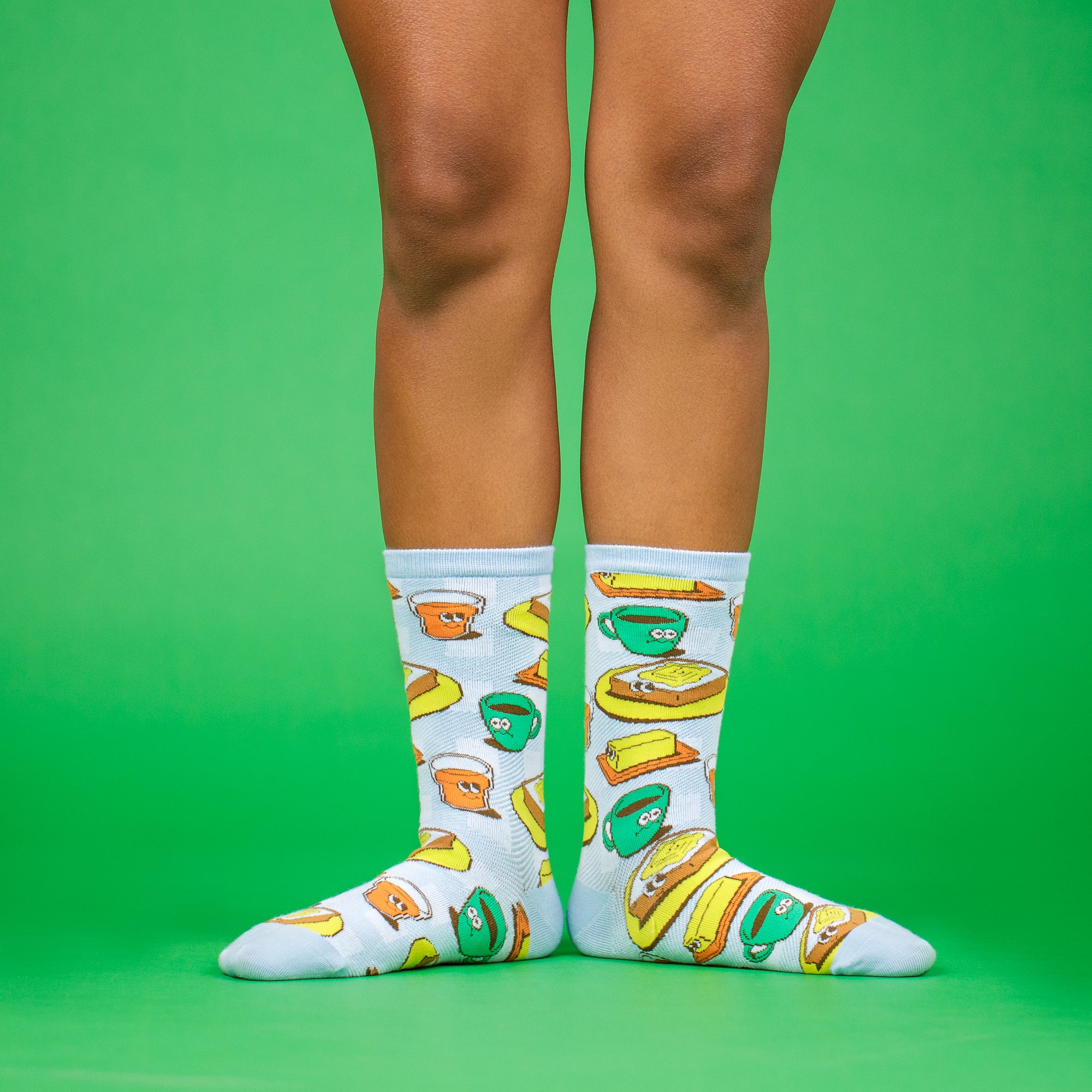 A green background showing two legs from the knee down in a ballet first position pose, wearing ankle socks from the Awesome Socks Club with colorful illustrations of toast, butter and coffee. 