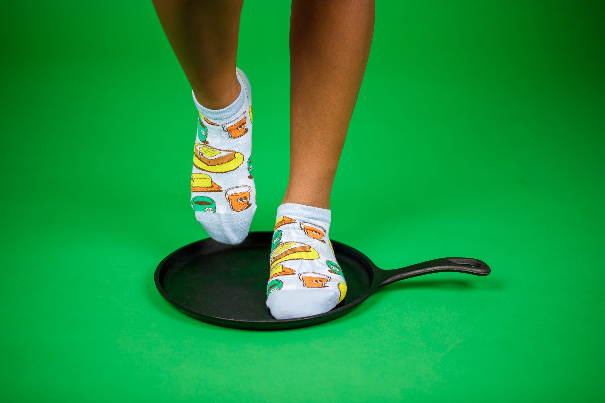 A green background showing two legs from the knee down, wearing ankle socks from the Awesome Socks Club with colorful illustrations of toast, butter and coffee. One of the feet is standing on a flat cast-iron pan.