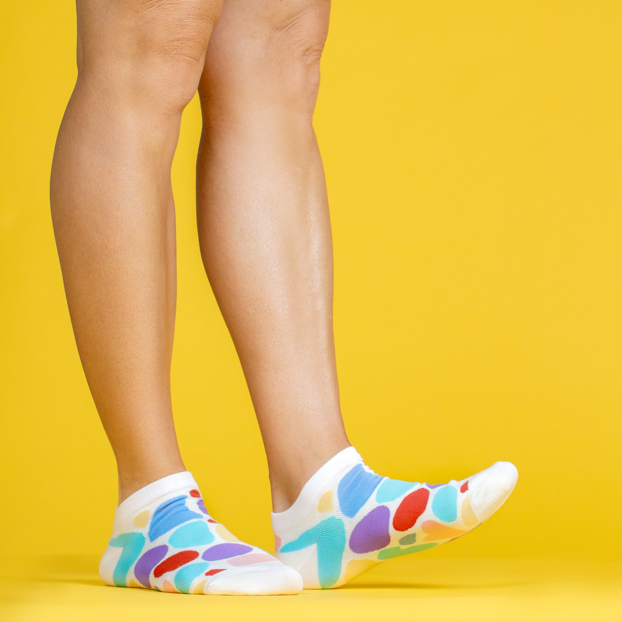 A yellow background showing two legs from the knee down, wearing ankle socks from the Awesome Socks Club with colorful blobs on them.