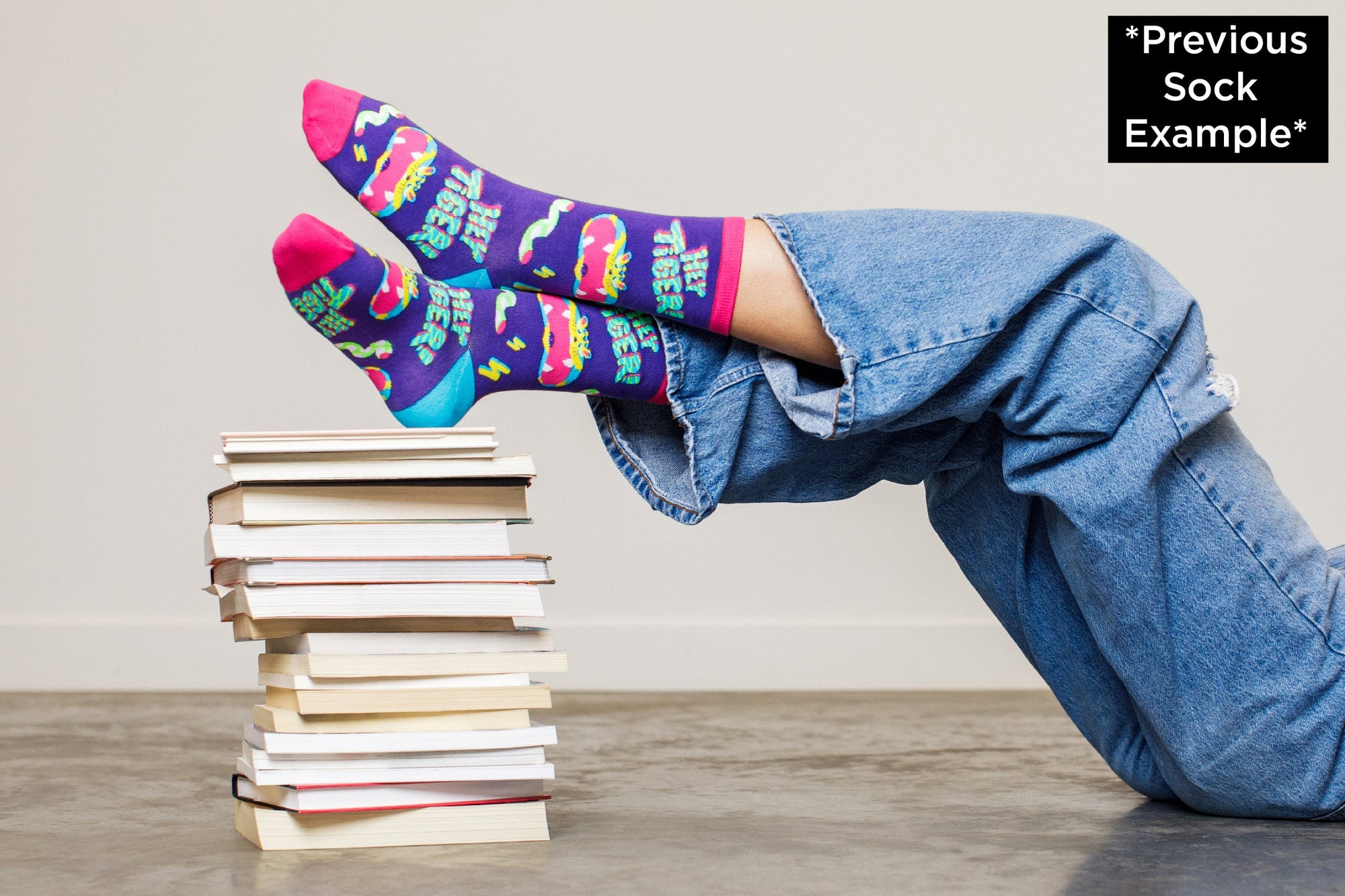 A pair of colorful socks from the Awesome Socks Club with an abstract tiger and the words "Hey Tiger". Socks are on the feet of someone laying down with their feet propped up on books.