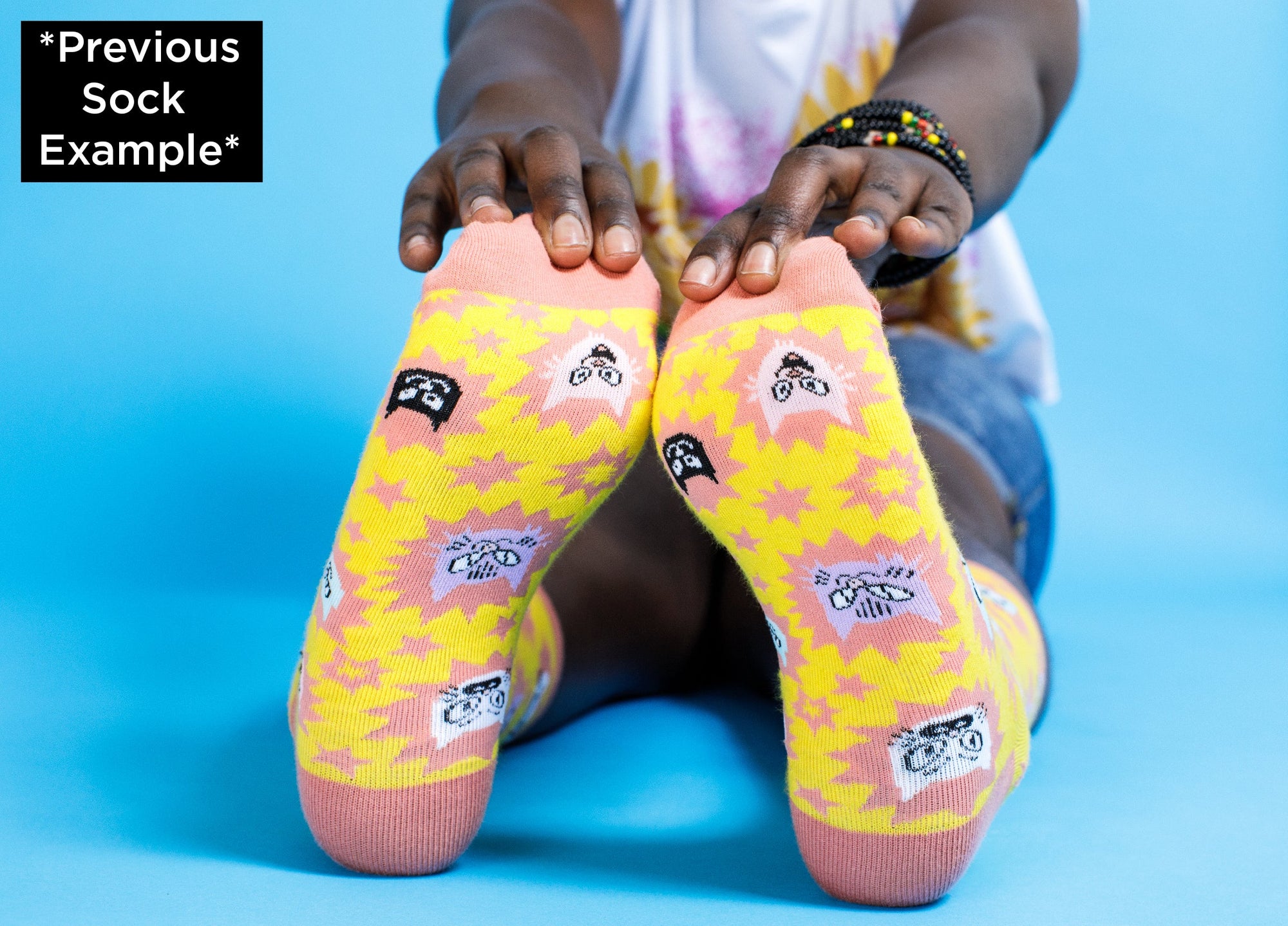 A pair of yellow and pink socks from the Awesome Socks Club with cat heads with different facial expressions. Socks are on the feet of someone sitting down and stretching, holding their feet.